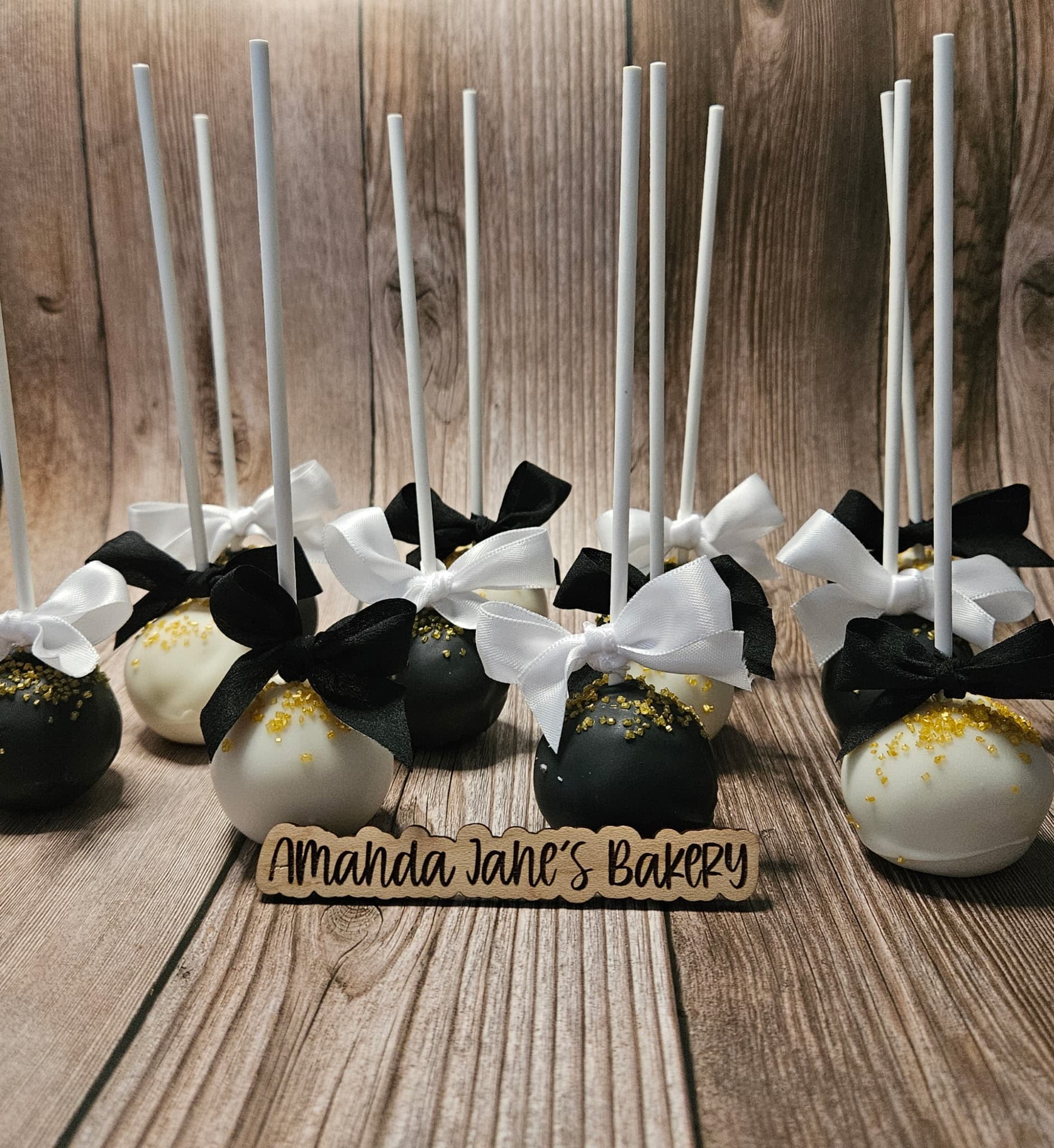 Black, white, and gold cake pops displayed beside an Amanda Jane's Bakery sign